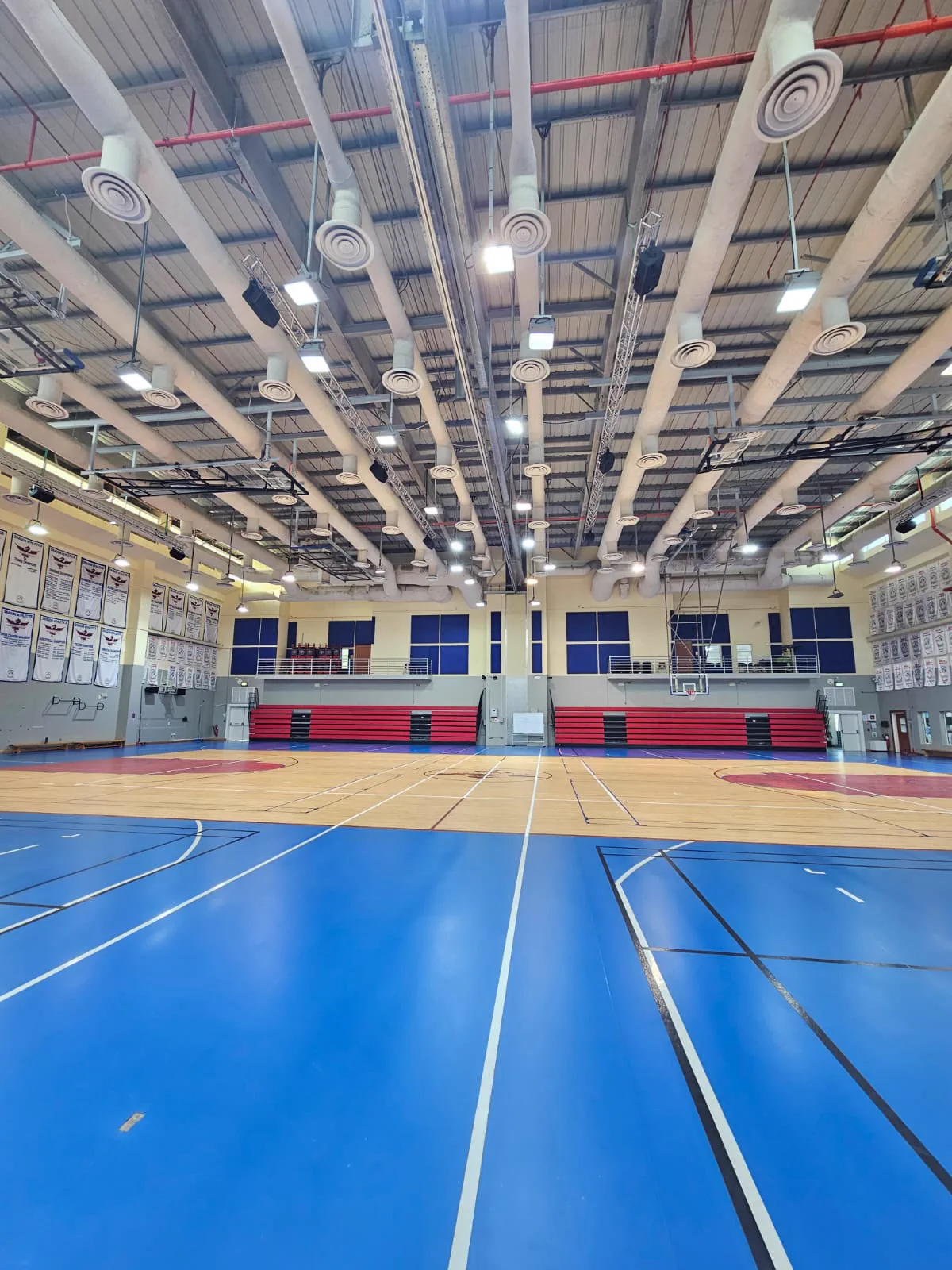 Interior of a school gymnasium in Doha showing acoustic ceiling baffles, high-bay lighting, maple court and red telescopic bleacher seating
